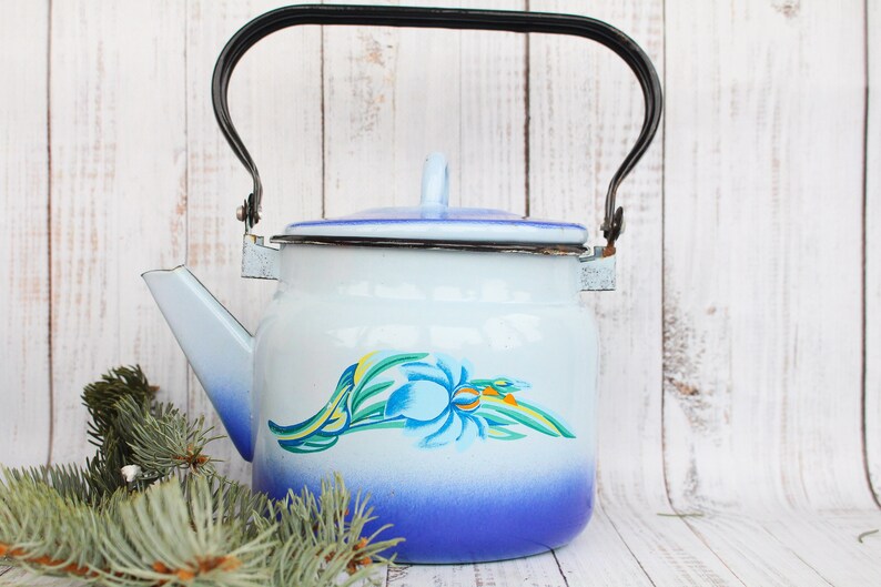 Op de afbeelding: Een blauwe en witte emaille theepot met een zwart handvat en een bloemenontwerp. De theepot staat op een wit houten oppervlak.