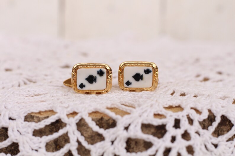 May include: A pair of gold cufflinks with a white background and black spades design.