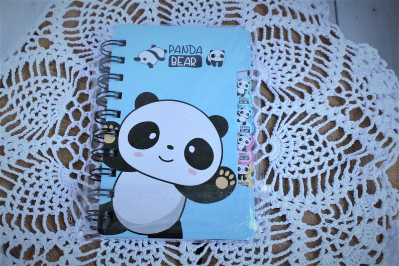 K&ouml;nnte beinhalten: Ein blaues Spiralnotizbuch mit einem Pandab&auml;r-Design. Der Einband zeigt einen gro&szlig;en Pandab&auml;r mit schwarzen Augen und einem wei&szlig;en Gesicht. Der Text "Panda Bear" ist in schwarzen Buchstaben auf dem Einband gedruckt.