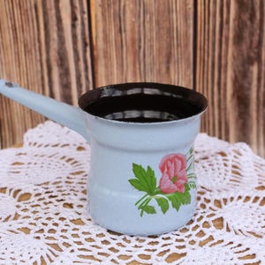 May include: A light blue enamel coffee pot with a black interior and a floral design featuring a pink flower and green leaves. The pot is sitting on a white crocheted doily.