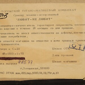 May include: A close-up of a paper tag with black text on a beige background. The tag is for a souvenir made by the Zaporozhye Titanium-Magnesium Combine. The text reads "Souvenir, made of titanium, "Loves - Doesn't Love." The tag also includes information about the manufacturer's warranty, the product's article number, and the date of manufacture.