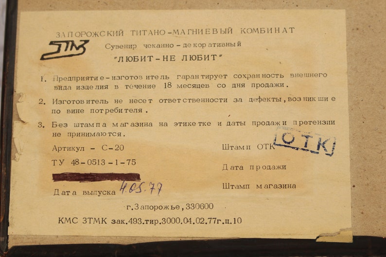 May include: A close-up of a paper tag with black text on a beige background. The tag is for a souvenir made by the Zaporozhye Titanium-Magnesium Combine. The text reads "Souvenir, made of titanium, "Loves - Doesn't Love." The tag also includes information about the manufacturer's warranty, the product's article number, and the date of manufacture.