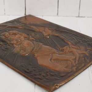 May include: A copper wall plaque with a raised relief design of a woman holding a child and a dog. The plaque has a dark brown patina and is set against a white background.