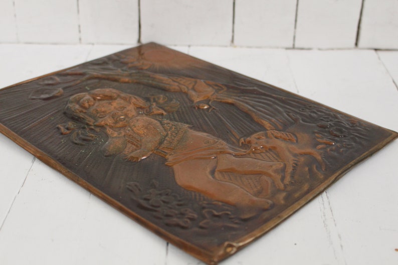 May include: A copper wall plaque with a raised relief design of a woman holding a child and a dog. The plaque has a dark brown patina and is set against a white background.