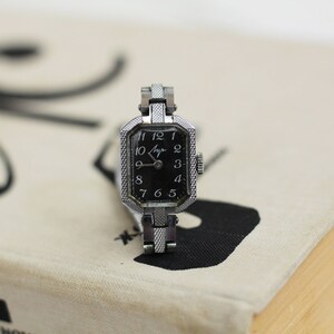K&ouml;nnte beinhalten: Eine silberne Armbanduhr mit schwarzem Zifferblatt und wei&szlig;en Ziffern. Die Uhr hat ein quadratisches Zifferblatt und ein silbernes Armband.