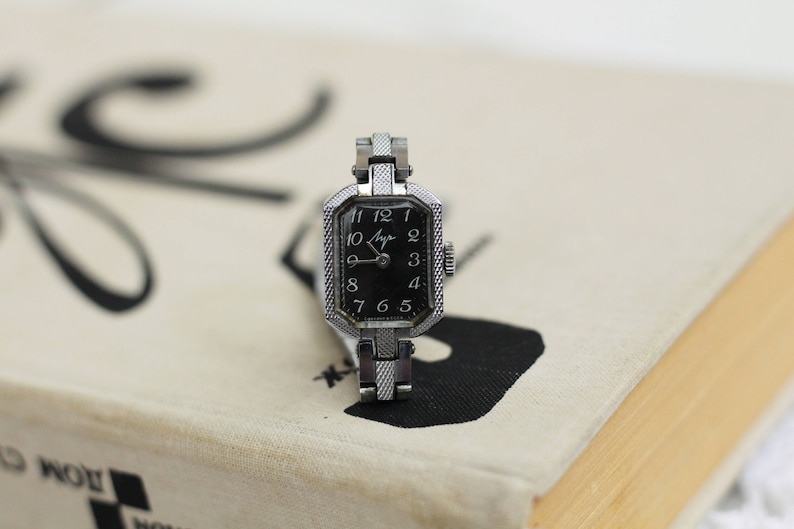 K&ouml;nnte beinhalten: Eine silberne Armbanduhr mit schwarzem Zifferblatt und wei&szlig;en Ziffern. Die Uhr hat ein quadratisches Zifferblatt und ein silbernes Armband.