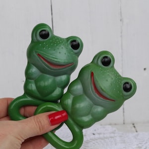 May include: A pair of green rubber frog rattles with smiling faces and black eyes. The frogs are connected by a green ring.