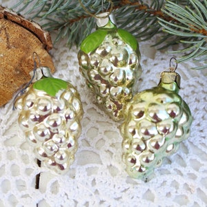 May include: Three vintage glass Christmas ornaments shaped like bunches of grapes. The ornaments are silver with green leaves and are hanging from a white crocheted doily.