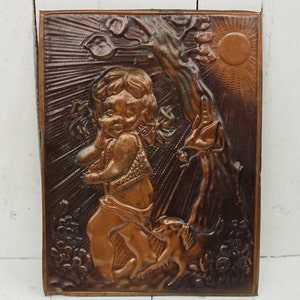 May include: A copper wall plaque with a raised relief design of a young girl with a dog. The girl is wearing a dress and has her hair in pigtails. The dog is sitting next to her and looking up at her. The plaque is set against a white wooden background.