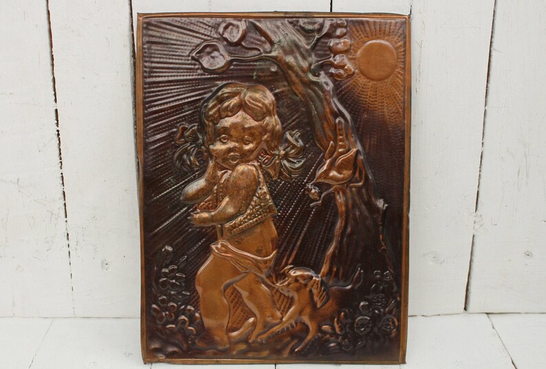 May include: A copper wall plaque with a raised relief design of a young girl with a dog. The girl is wearing a dress and has her hair in pigtails. The dog is sitting next to her and looking up at her. The plaque is set against a white wooden background.