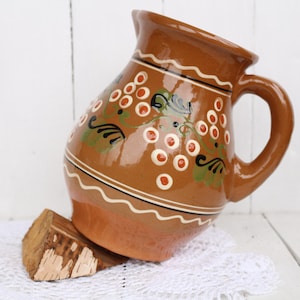 Puede incluir: Una jarra de cerámica marrón con un diseño floral blanco y verde. La jarra tiene un asa y una boca ancha.