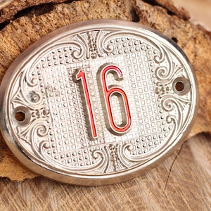 May include: A silver oval-shaped metal plaque with an ornate design and the number 16 in red lettering.