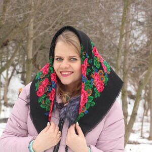 Op de afbeelding: Een vrouw draagt een zwarte hoofddoek met een bloemenpatroon in tinten rood, groen en blauw. Ze glimlacht en kijkt naar de camera. De achtergrond is een wazig beeld van een besneeuwd bos.