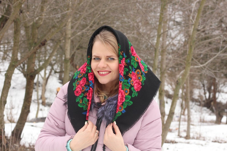 Op de afbeelding: Een vrouw draagt een zwarte hoofddoek met een bloemenpatroon in tinten rood, groen en blauw. Ze glimlacht en kijkt naar de camera. De achtergrond is een wazig beeld van een besneeuwd bos.