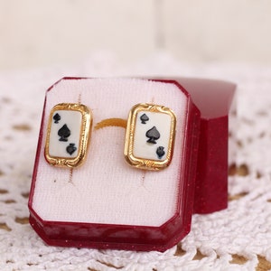 May include: A pair of gold earrings with a white background and black spades. The earrings are in a red box.