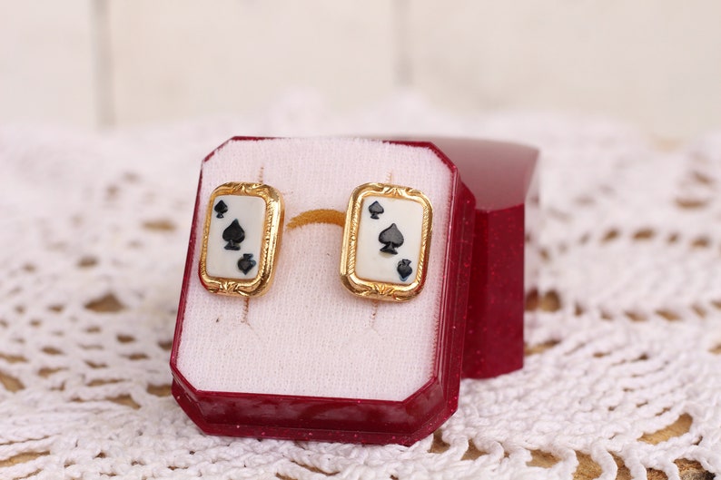 May include: A pair of gold earrings with a white background and black spades. The earrings are in a red box.