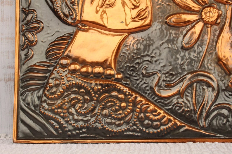 May include: A copper wall plaque with a raised design of a woman's face and a flower. The plaque has a dark brown patina and is embossed with intricate details.