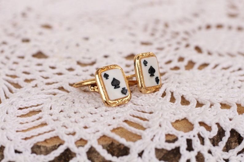May include: A pair of gold cufflinks with a white enamel background and black spade suit design.