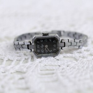 K&ouml;nnte beinhalten: Eine silberne Armbanduhr mit schwarzem Zifferblatt und silbernem Metallband. Das Zifferblatt der Uhr hat schwarze r&ouml;mische Ziffern als Stundenmarkierungen und einen silbernen Sekundenzeiger.
