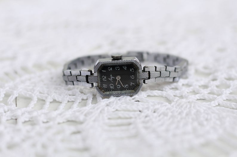 K&ouml;nnte beinhalten: Eine silberne Armbanduhr mit schwarzem Zifferblatt und silbernem Metallband. Das Zifferblatt der Uhr hat schwarze r&ouml;mische Ziffern als Stundenmarkierungen und einen silbernen Sekundenzeiger.
