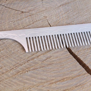 Könnte beinhalten: Ein silberner Kamm mit einem dekorativen Design am Griff. Der Kamm hat feine Zähne und ist aus Metall.