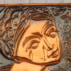 May include: A copper wall plaque with a raised relief design of a woman's face. The woman's face is surrounded by floral designs.