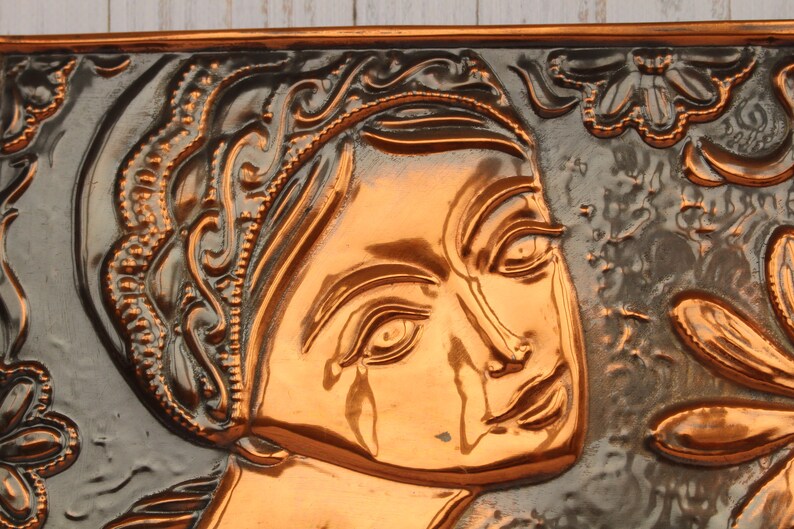 May include: A copper wall plaque with a raised relief design of a woman's face. The woman's face is surrounded by floral designs.