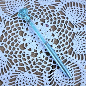 K&ouml;nnte beinhalten: Ein hellblauer Stift mit einem gro&szlig;en, klaren, facettierten, blauen Stein an der Spitze. Der Stift ist mit lockerem, hellblauem Glitzer gef&uuml;llt.