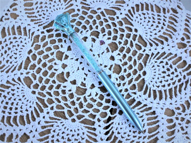 K&ouml;nnte beinhalten: Ein hellblauer Stift mit einem gro&szlig;en, klaren, facettierten, blauen Stein an der Spitze. Der Stift ist mit lockerem, hellblauem Glitzer gef&uuml;llt.