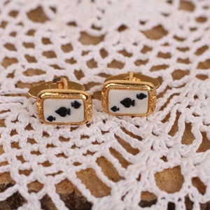 May include: A pair of gold cufflinks with a white background and black spade playing card symbols.
