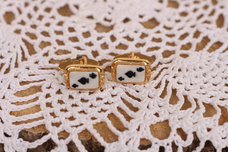 May include: A pair of gold cufflinks with a white background and black spade playing card symbols.