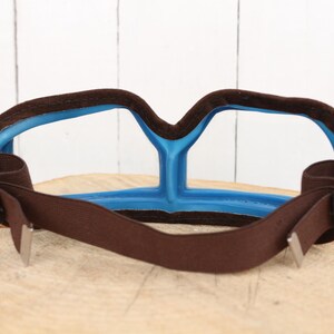 May include: Brown and blue safety goggles with an adjustable strap. The goggles have a clear lens and a soft, padded frame.