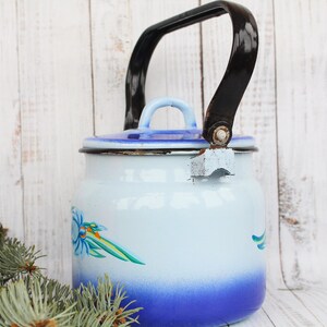 Op de afbeelding: Een blauwe en witte emaille theepot met een zwart handvat en een bloemenontwerp. De theepot staat op een wit houten oppervlak.