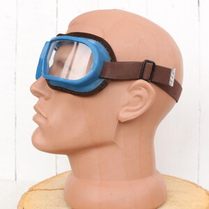 May include: Blue safety goggles with a brown adjustable strap. The goggles have a clear lens and are worn on a mannequin head.