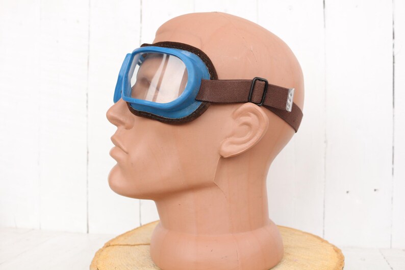 May include: Blue safety goggles with a brown adjustable strap. The goggles have a clear lens and are worn on a mannequin head.