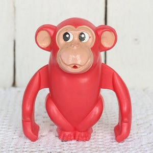 May include: A bright red plastic monkey toy with large, round eyes and a wide smile. The monkey has long arms and legs, and its body is shaped like a rounded rectangle.