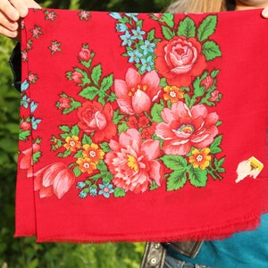 以下が含まれることがあります： ピンク、黄色、青の花が咲いた花柄の赤い布のストール。ストールはフリンジの縁取りがされている。