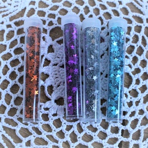K&ouml;nnte beinhalten: Vier kleine, durchsichtige R&ouml;hrchen gef&uuml;llt mit sternf&ouml;rmigem Glitter. Der Glitter ist in vier Farben: orange, lila, silber und blau.