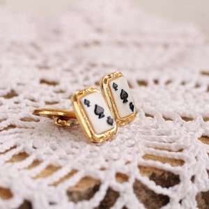 May include: A pair of gold cufflinks with a white background and black spades. The cufflinks are on a white crocheted surface.