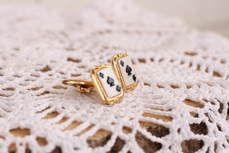 May include: A pair of gold cufflinks with a white background and black spades. The cufflinks are on a white crocheted surface.