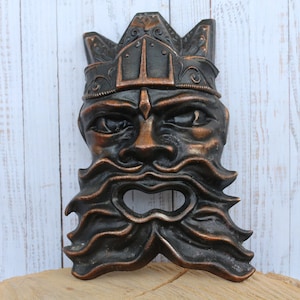 May include: A dark brown and copper colored metal mask with a crown, beard, and a hole in the mouth. The mask is on a wooden surface.