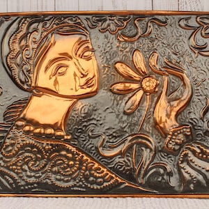 May include: A copper wall sculpture depicting a woman with a tear on her cheek, holding a flower. The sculpture is embossed with intricate details and has a dark gray background.