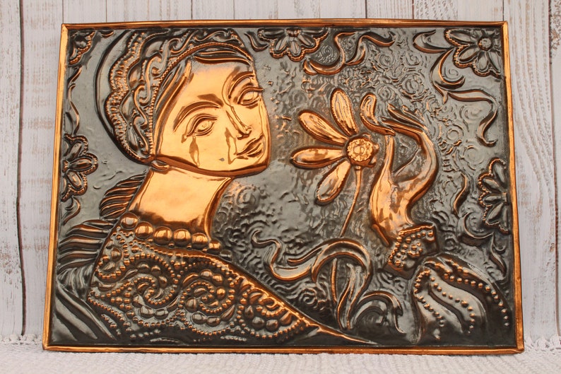 May include: A copper wall sculpture depicting a woman with a tear on her cheek, holding a flower. The sculpture is embossed with intricate details and has a dark gray background.