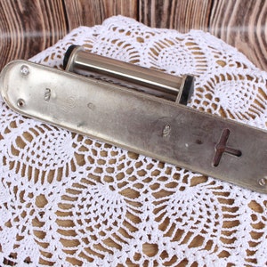 K&ouml;nnte beinhalten: Eine silberfarbene Metallrolle mit einem schwarzen Knopf an jedem Ende, die auf einer wei&szlig;en H&auml;kelspitze liegt. Die Rolle ist an einer Metallplatte mit zwei L&ouml;chern und einer kreuzf&ouml;rmigen &Ouml;ffnung befestigt.