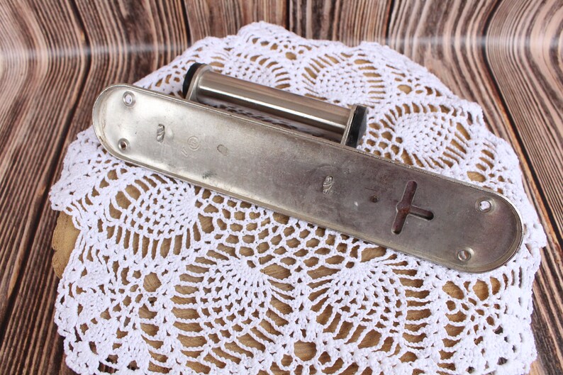 K&ouml;nnte beinhalten: Eine silberfarbene Metallrolle mit einem schwarzen Knopf an jedem Ende, die auf einer wei&szlig;en H&auml;kelspitze liegt. Die Rolle ist an einer Metallplatte mit zwei L&ouml;chern und einer kreuzf&ouml;rmigen &Ouml;ffnung befestigt.