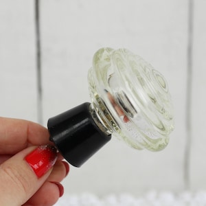 May include: A clear glass bottle stopper with a black plastic base. The glass is layered and has a rounded shape.