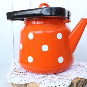 Könnte beinhalten: Eine orangefarbene Emaille-Teekanne mit weißen Tupfen und einem schwarzen Griff und Deckel. Die Teekanne hat einen Ausguss und einen kleinen orangefarbenen Griff am Deckel. Sie steht auf einer weißen Spitzendecke auf einer Holzscheibe.