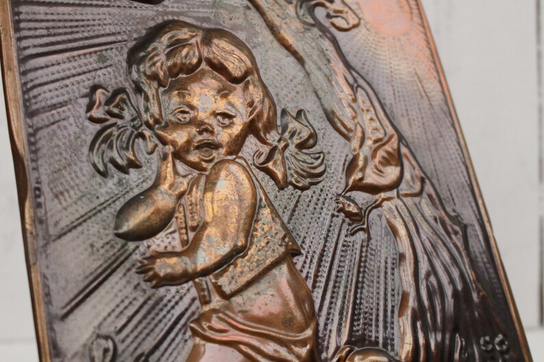 May include: A copper wall plaque with a raised relief design of a young girl with pigtails, wearing a dress and holding a doll. The girl is looking to the right and has a surprised expression on her face. The background is a textured copper surface with a sunburst pattern.