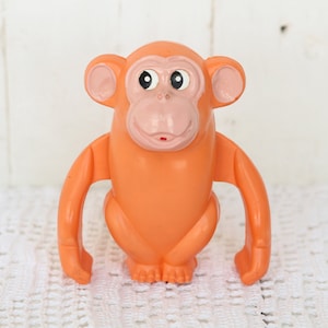 May include: Orange plastic toy monkey with large eyes and pink ears. The monkey has a wide smile and is standing on a white surface.