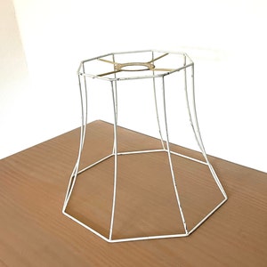 May include: A white metal lampshade frame with an octagonal top and base. The frame has a slightly flared shape and is made of thin, painted metal rods. The frame sits on a brown wooden surface.